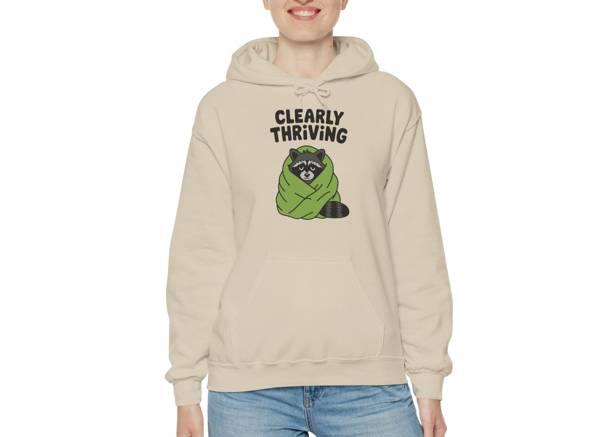 Model wearing a beige hoodie featuring the text "Clearly Thriving" and a cartoon illustration of a raccoon wrapped in a cozy blanket. Part of the Vivid Divergence Sensory Friendly Unisex Hoodie Range.