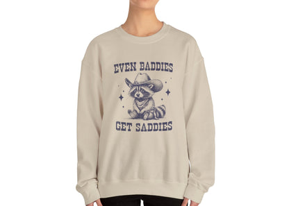 Model wearing a beige sweater jumper with the text "Even Baddies Get Saddies" and a cartoon raccoon illustration. Part of the Vivid Divergence Sensory Friendly Unisex Sweater Jumper Range.