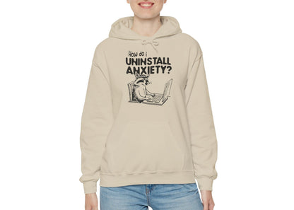 Model wearing a beige hoodie featuring the text "How Do I Uninstall Anxiety?" and a cartoon illustration of a raccoon using a laptop. Part of the Vivid Divergence Sensory Friendly Unisex Hoodie Range. 