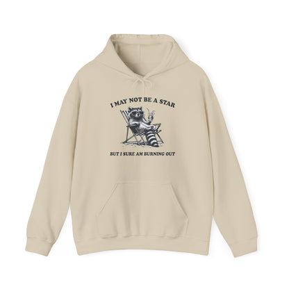 Front view of a beige hoodie featuring a raccoon sitting in a trash can with the text "I May Not Be A Star, But I Sure Am Burning Out." Part of the Vivid Divergence Sensory Friendly Unisex Hoodie Range.