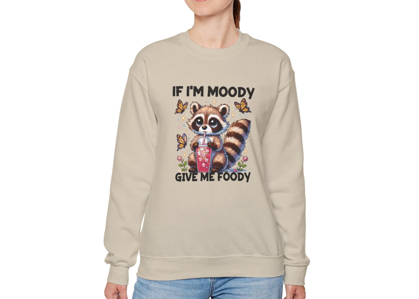 Model wearing a beige sweater jumper with the text "If I'm Moody, Give Me Foody" and a cartoon raccoon illustration holding snacks. Part of the Vivid Divergence Sensory Friendly Unisex Sweater Jumper Range.