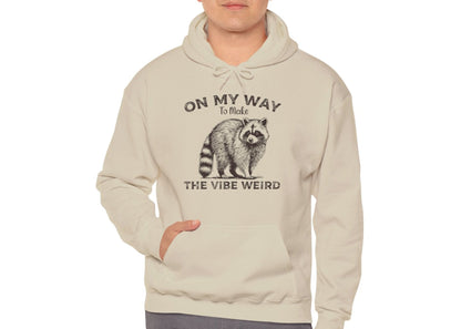 Model wearing a light blue hoodie featuring the text "On My Way to Make the Vibe Weird" and a cartoon illustration of a raccoon holding a stick. Part of the Vivid Divergence Sensory Friendly Unisex Hoodie Range.