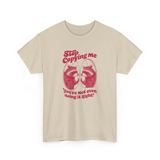 Front view of a beige T-shirt with "Stop Copying Me" in bold red text and a mirrored raccoon graphic, featuring the tagline "You're Not Even Doing It Right!" Part of the Vivid Divergence Sensory Friendly Unisex Tees Range.
