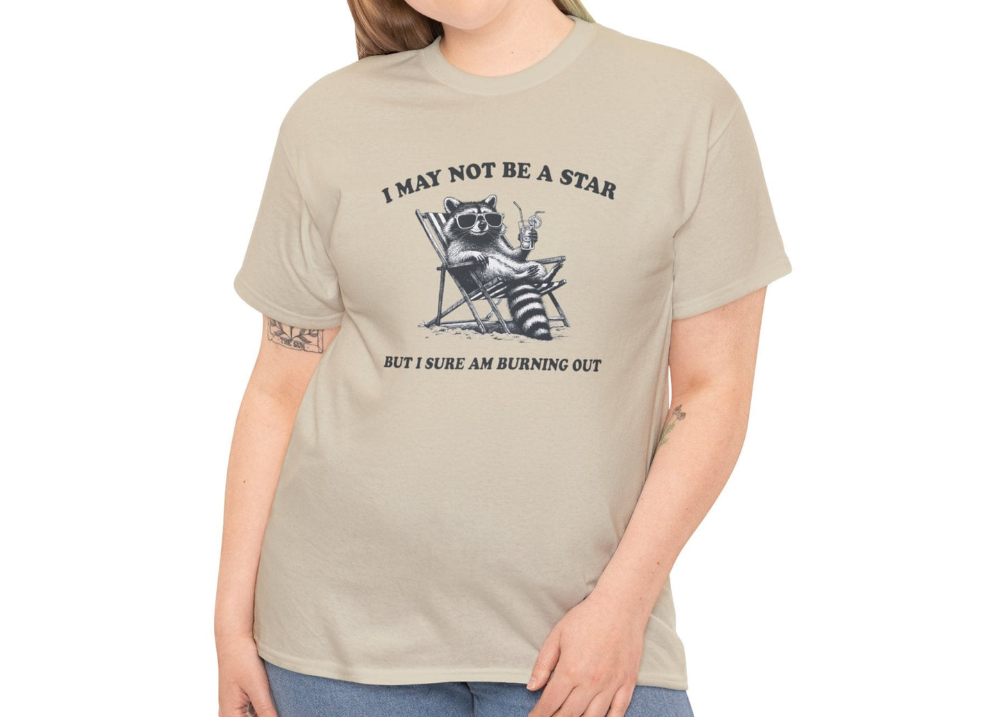 Modeled front view of a beige T-shirt featuring a cute raccoon holding a star with text that reads "I May Not Be A Star, But I Sure Am Burning Out." Part of the Vivid Divergence Sensory Friendly Unisex Tees Range.