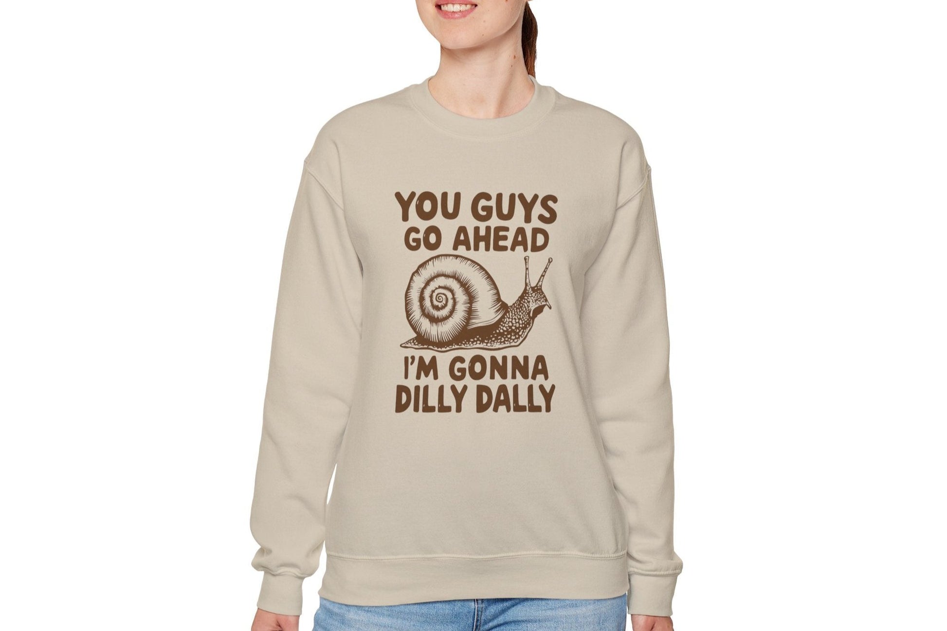 Model wearing a beige sweater jumper featuring the text "You Guys Go Ahead, I’m Gonna Dilly Dally" and a cartoon snail illustration. Part of the Vivid Divergence Sensory Friendly Unisex Sweater Jumper Range.