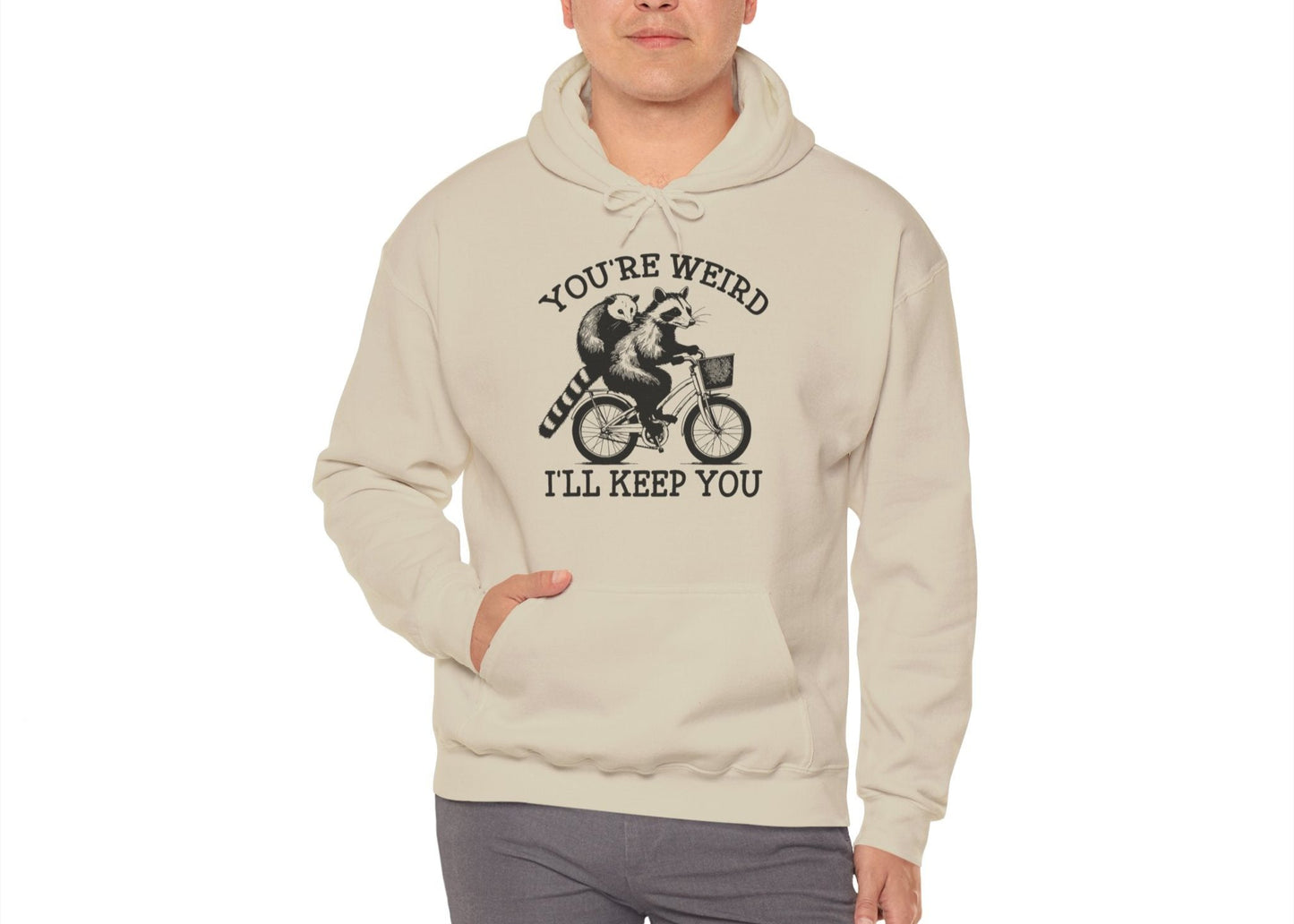 Model wearing a beige hoodie featuring the text "You're Weird, I'll Keep You" with a cartoon illustration of two raccoons riding a bicycle. Part of the Vivid Divergence Sensory Friendly Unisex Hoodie Range.
