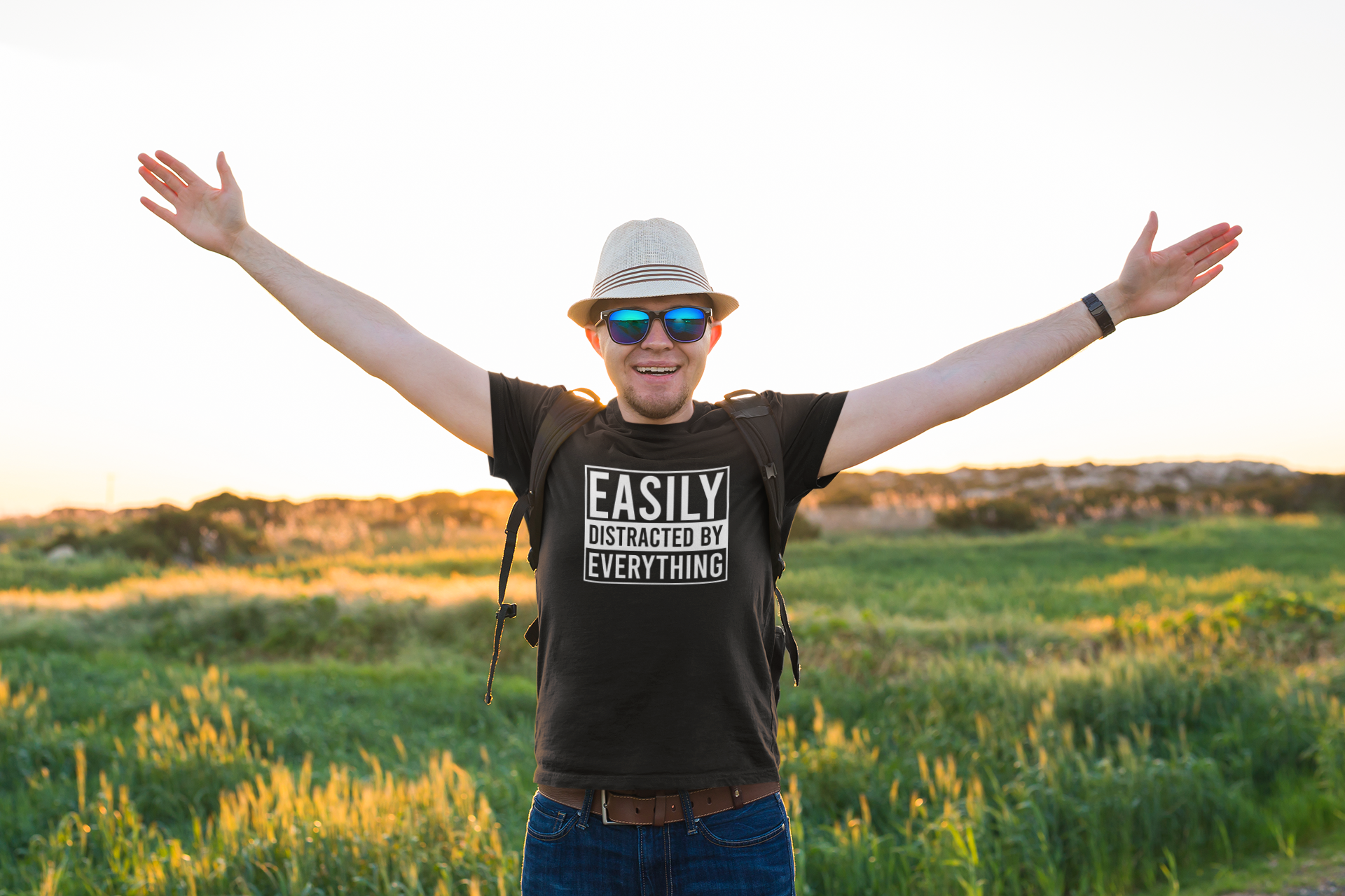 A person outdoors wearing a black T-shirt with the text "Easily Distracted by Everything," arms raised against a field background. Part of the Vivid Divergence Sensory Friendly Unisex Tees Range.