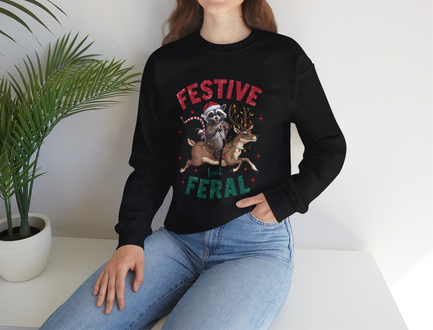 Lifestyle image of a black sweatshirt with a raccoon holding Christmas lights and the text "Festive but Feral," styled in a cozy indoor environment. Part of the Vivid Divergence Sensory Friendly Unisex Sweater Jumper Range.