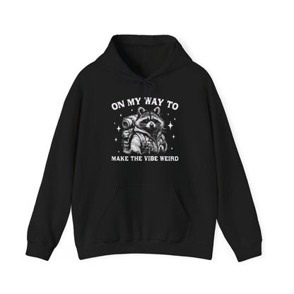 Front view of a black hoodie featuring the text "On My Way to Make the Vibe Weird" and a cartoon illustration of a raccoon holding a stick. Part of the Vivid Divergence Sensory Friendly Unisex Hoodie Range.