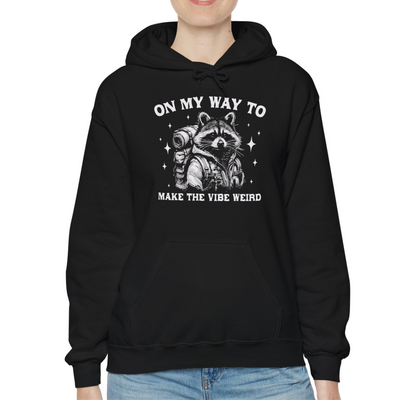 Model wearing a black hoodie featuring the text "On My Way to Make the Vibe Weird" and a cartoon illustration of a raccoon holding a stick. Part of the Vivid Divergence Sensory Friendly Unisex Hoodie Range.