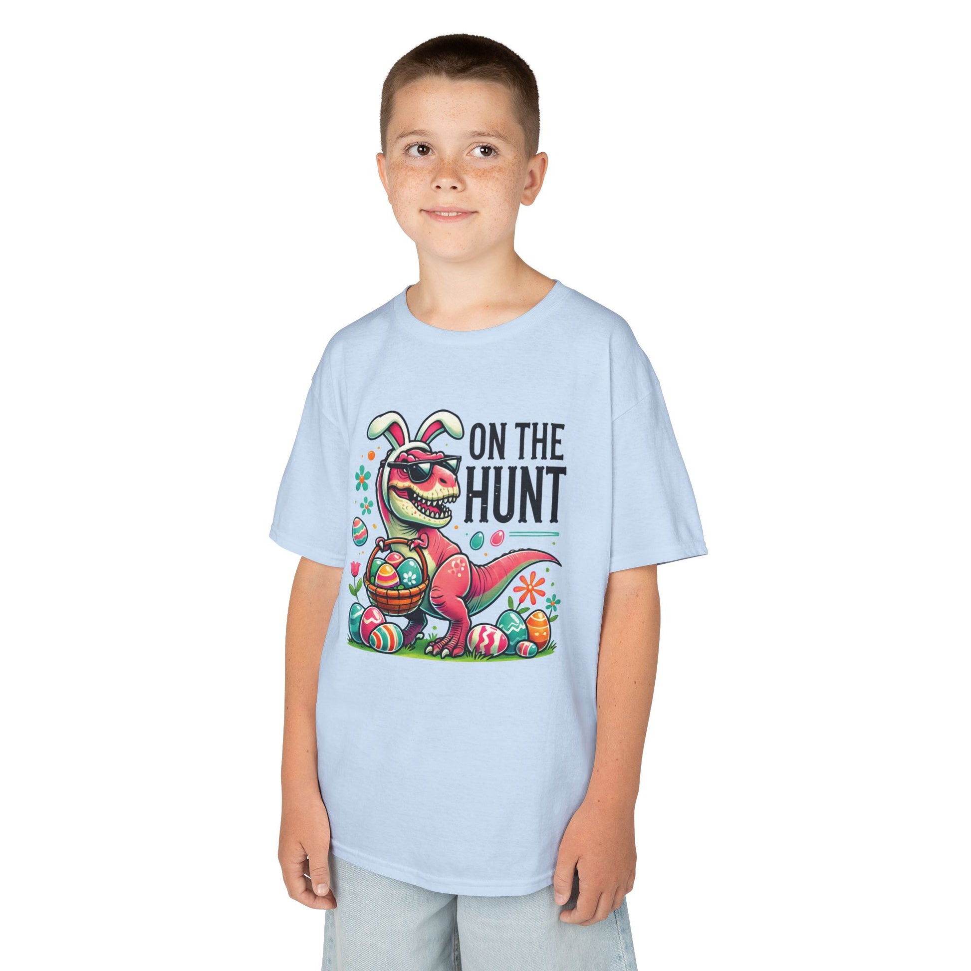 Child wearing a light blue Easter T-shirt featuring a dinosaur, "On the Hunt" text, and colorful eggs. A fun pick for Easter egg hunts. Hop into the season in style and shop Easter shirts online today!
