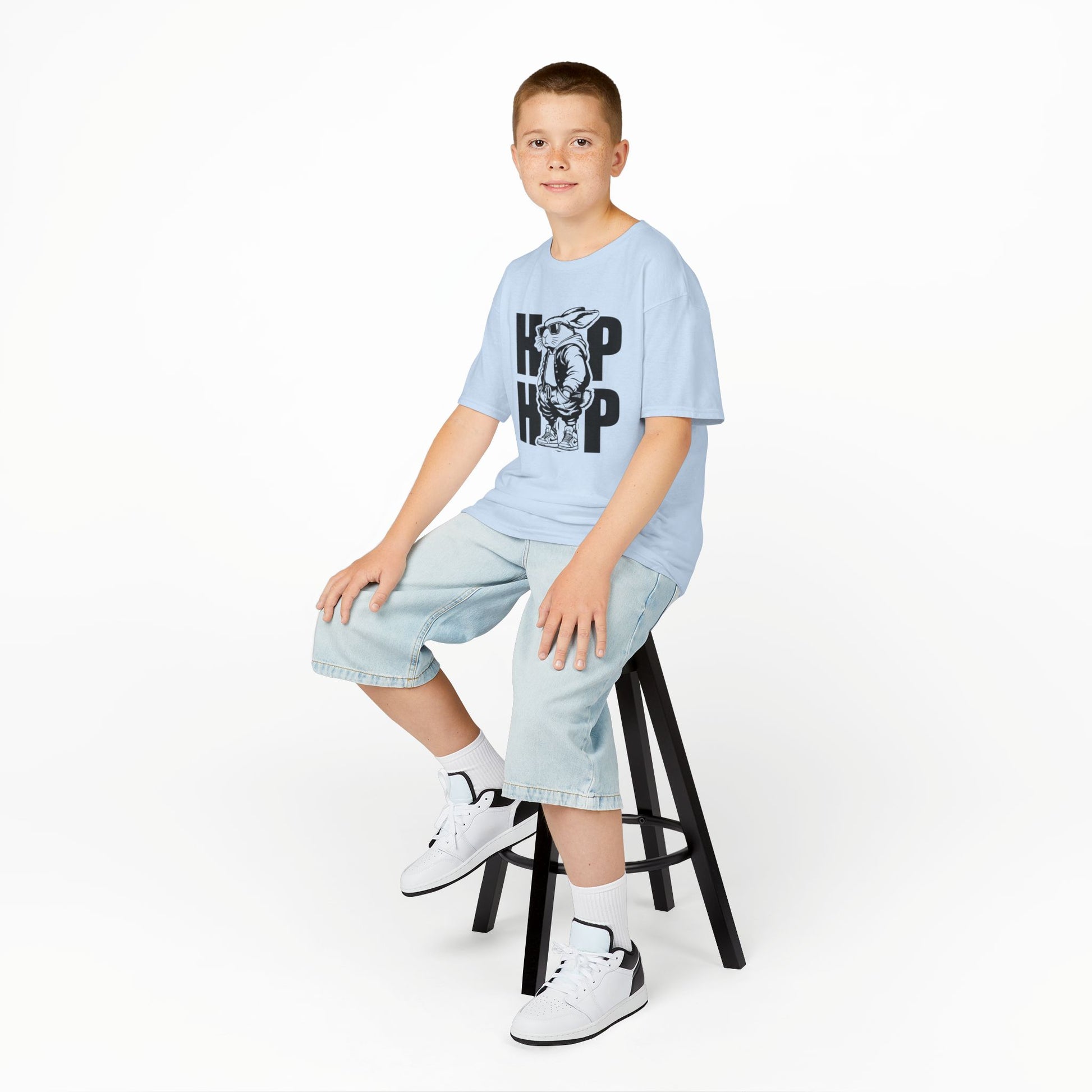 Child wearing a light blue hip-hop bunny Easter T-shirt while sitting on a stool. A stylish pick for Easter celebrations. Hop into the season in style and shop Easter shirts online today!