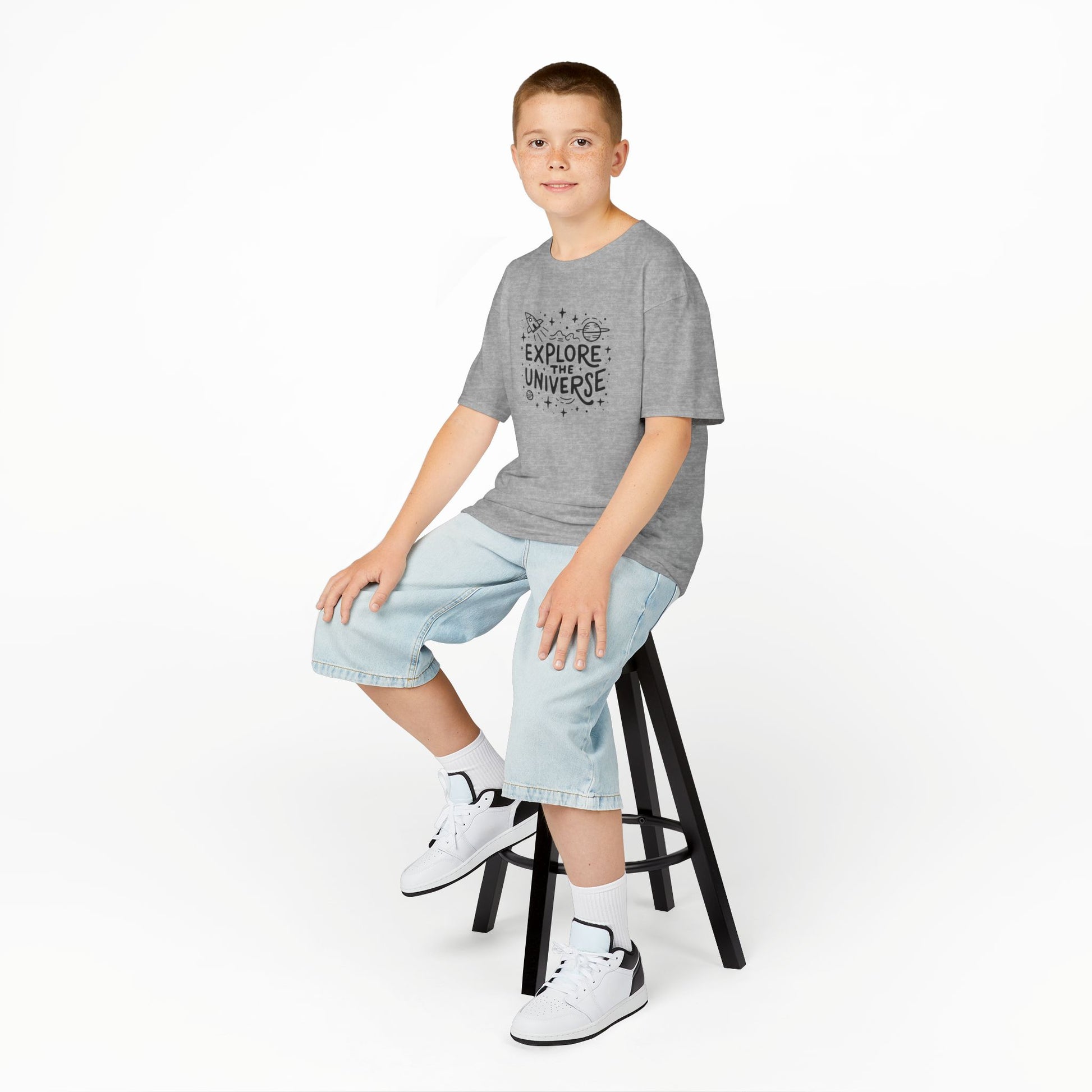 Boy sitting on a stool wearing a gray "Explore The Universe" kids T-shirt with a space-themed graphic. Part of the Vivid Divergence Sensory Friendly Kids Shirts Range. The perfect T-shirt for sensory-sensitive kids and comes with a Sensory Satisfaction Promise.