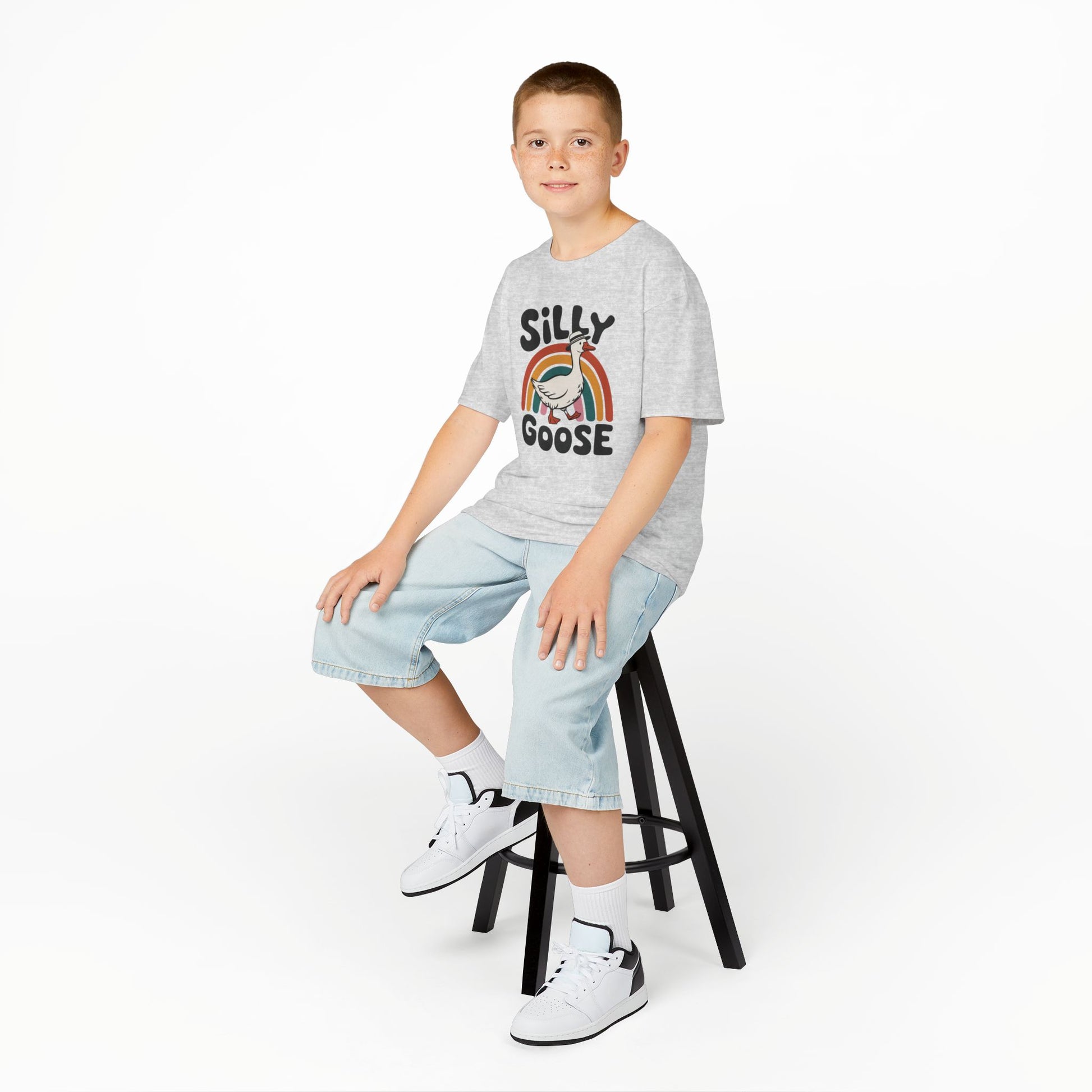 Boy sitting on a stool wearing a gray "Silly Goose" rainbow kids T-shirt with a cartoon goose and colorful rainbow. Part of the Vivid Divergence Sensory Friendly Kids Shirts Range. The perfect T-shirt for sensory-sensitive kids and comes with a Sensory Satisfaction Promise.