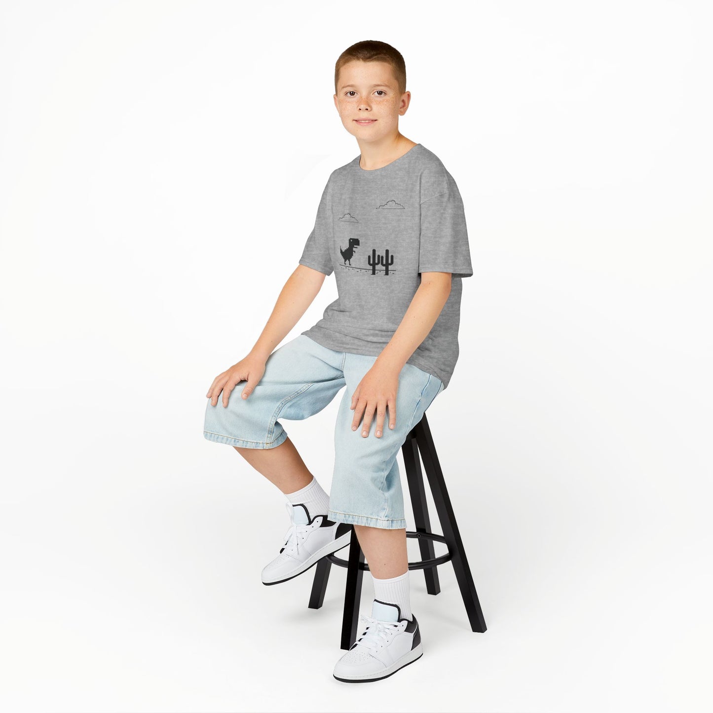 Boy sitting on a stool wearing a gray "T-Rex Runner" Dinosaur Game kids T-shirt with a pixelated T-Rex and cactus design. Part of the Vivid Divergence Sensory Friendly Kids Shirts Range. The perfect T-shirt for sensory-sensitive kids and comes with a Sensory Satisfaction Promise.