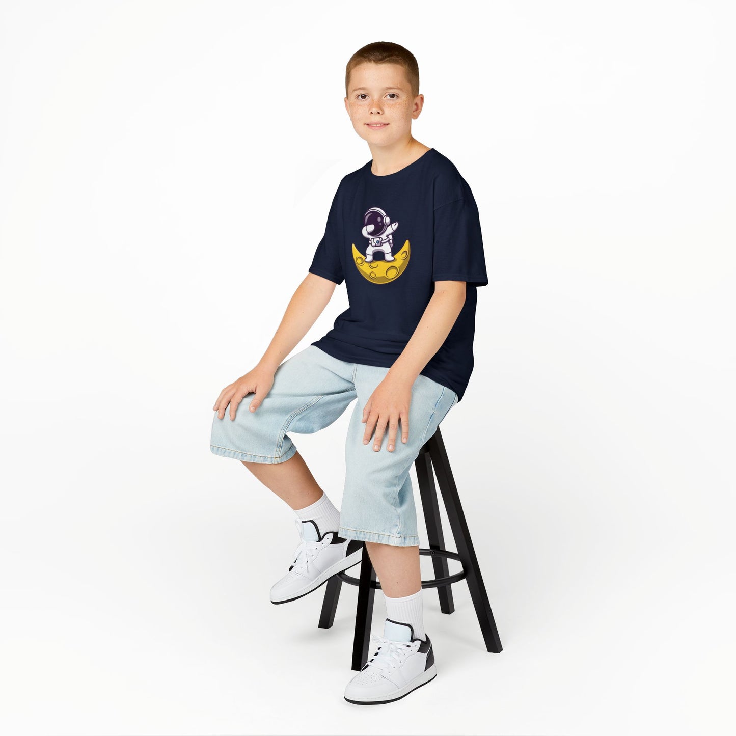 Boy sitting on a stool wearing a navy blue "Dabbing Astronaut on the Moon" kids T-shirt with an astronaut dabbing while sitting on a crescent moon. Part of the Vivid Divergence Sensory Friendly Kids Shirts Range. The perfect T-shirt for sensory-sensitive kids and comes with a Sensory Satisfaction Promise.