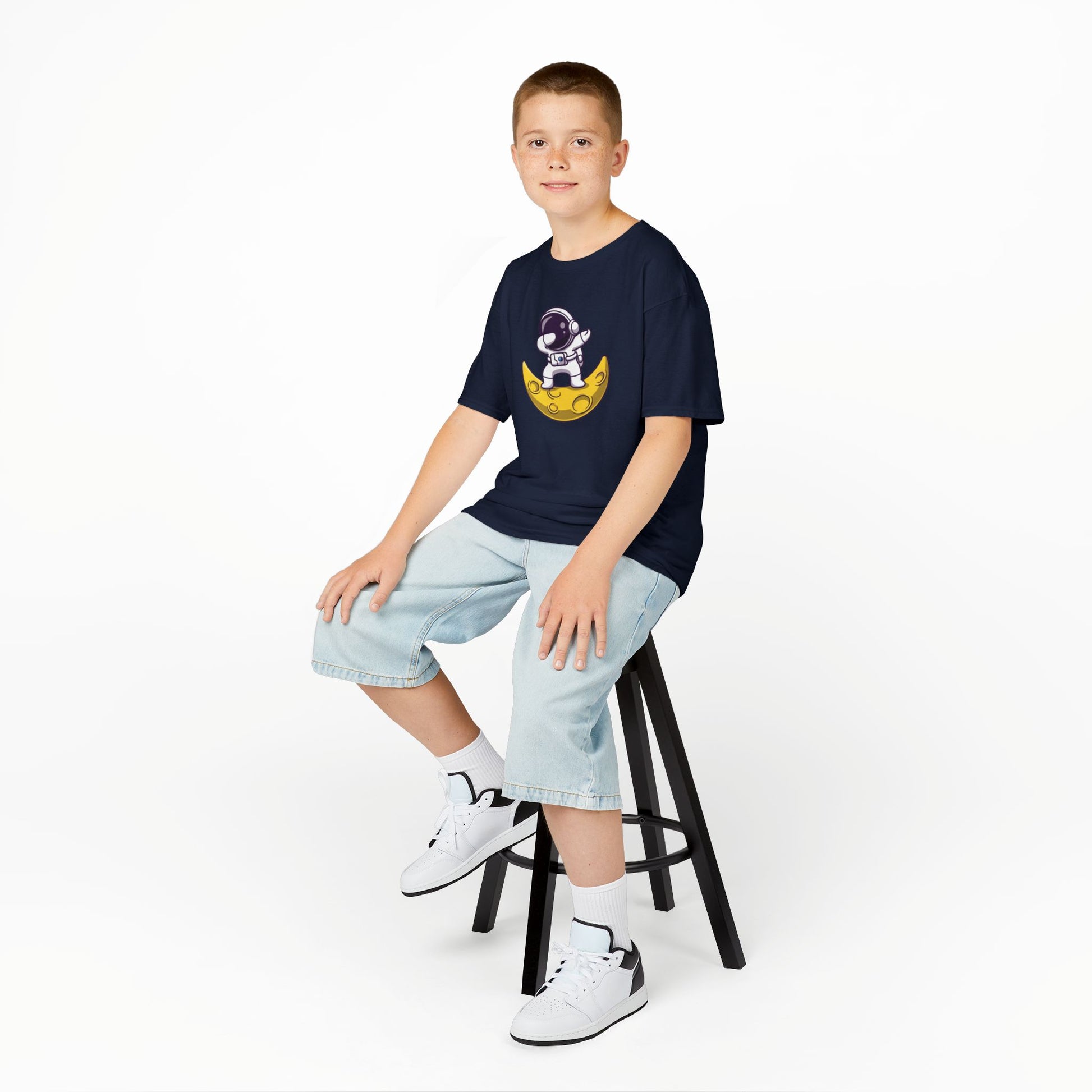 Boy sitting on a stool wearing a navy blue "Dabbing Astronaut on the Moon" kids T-shirt with an astronaut dabbing while sitting on a crescent moon. Part of the Vivid Divergence Sensory Friendly Kids Shirts Range. The perfect T-shirt for sensory-sensitive kids and comes with a Sensory Satisfaction Promise.