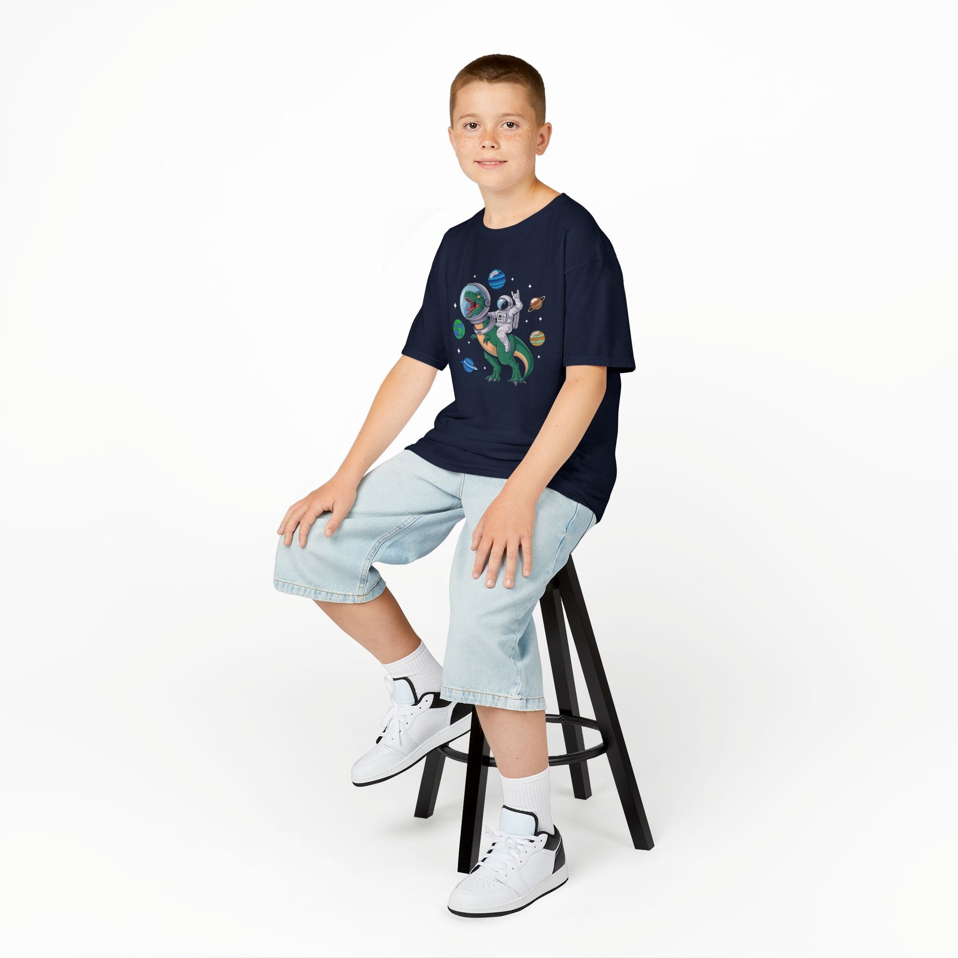 Boy sitting on a stool wearing a navy blue "Jurassic Space Jam" kids T-shirt with a dinosaur and astronaut floating among planets. Part of the Vivid Divergence Sensory Friendly Kids Shirts Range. The perfect T-shirt for sensory-sensitive kids and comes with a Sensory Satisfaction Promise.