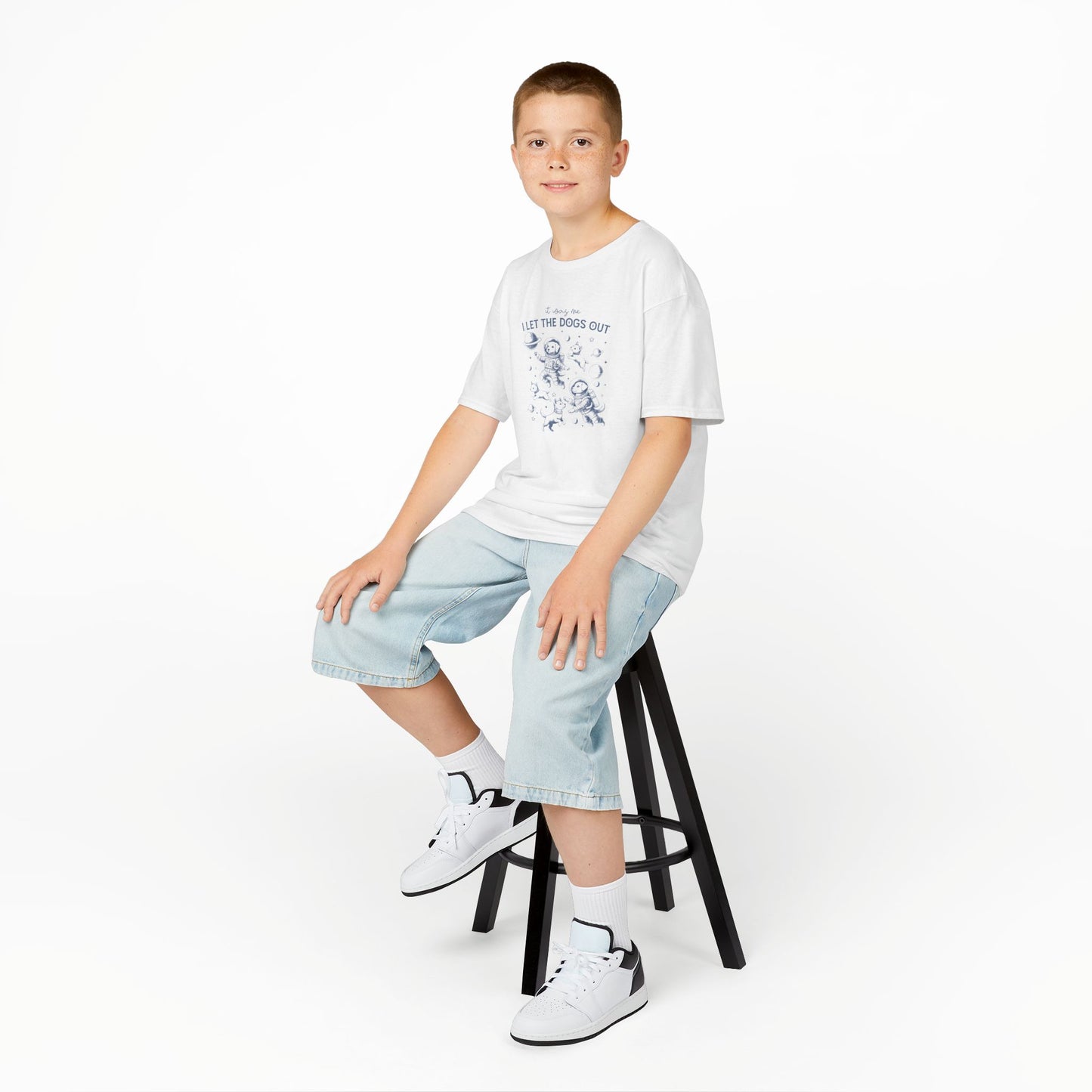 Boy sitting on a stool wearing a white "It Was Me, I Let The Dogs Out" kids T-shirt with playful dog graphics. Part of the Vivid Divergence Sensory Friendly Kids Shirts Range. The perfect T-shirt for sensory-sensitive kids and comes with a Sensory Satisfaction Promise.