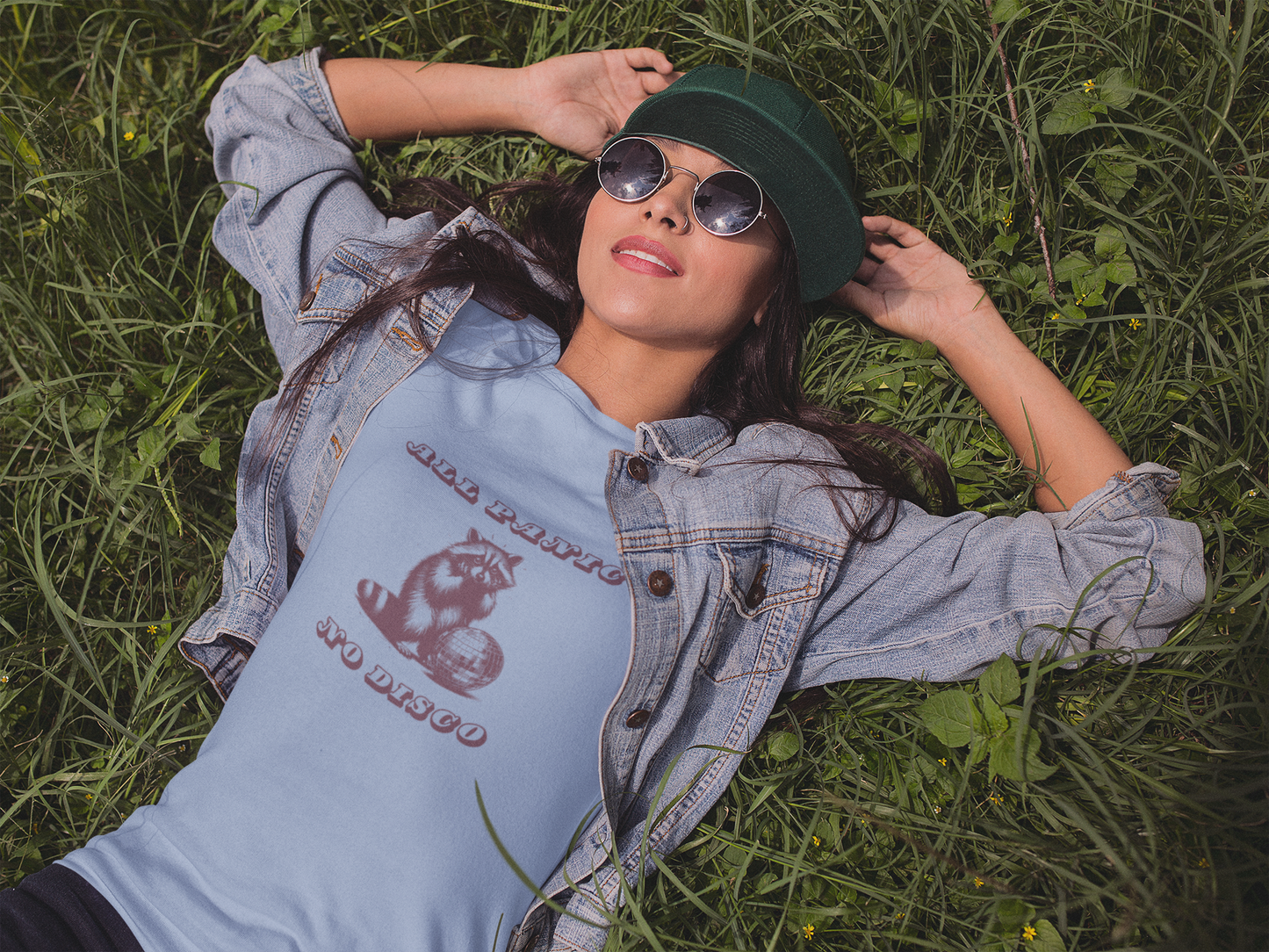Person lying on grass wearing a light blue T-shirt with the text "All Panic No Disco" and a raccoon illustration. Part of the Vivid Divergence Sensory Friendly Unisex Tees Range.