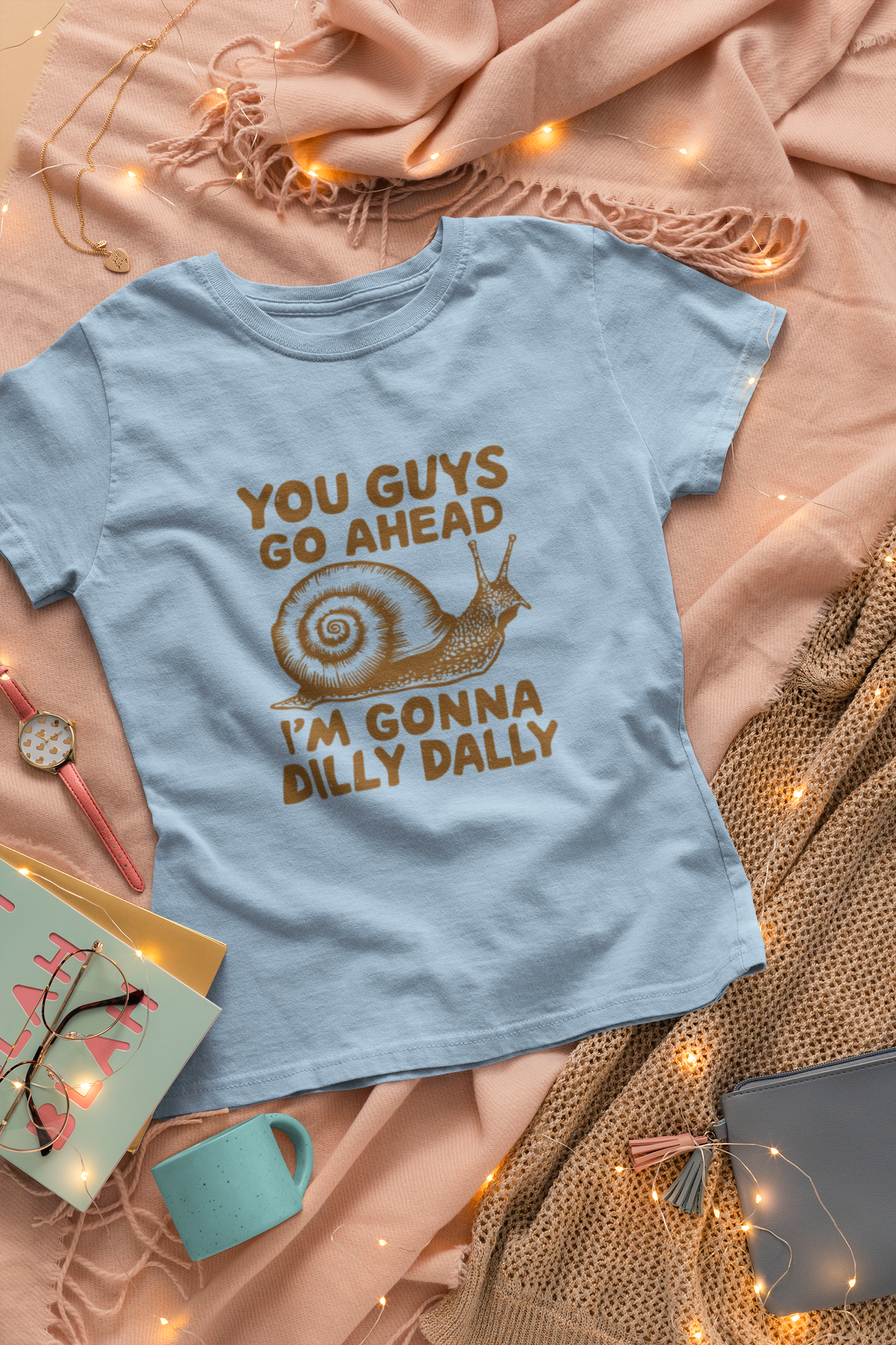Close-up view of a light blue T-shirt with the text "You Guys Go Ahead, I'm Gonna Dilly Dally" and a playful snail graphic in brown. Part of the Vivid Divergence Sensory Friendly Unisex Tees Range.
