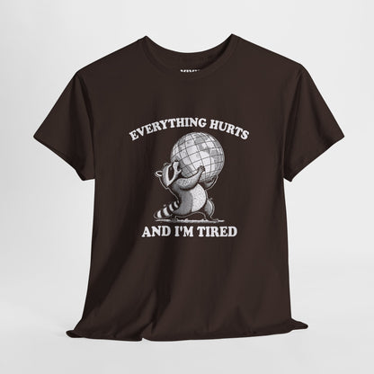 Hanging view of chocolate brown T-shirt featuring a vintage style sketch of a raccoon carrying the world with the phrase “Everything Hurts and I’m Tired.” The official uniform of burnt out. Part of the Vivid Divergence Memewear Collection!