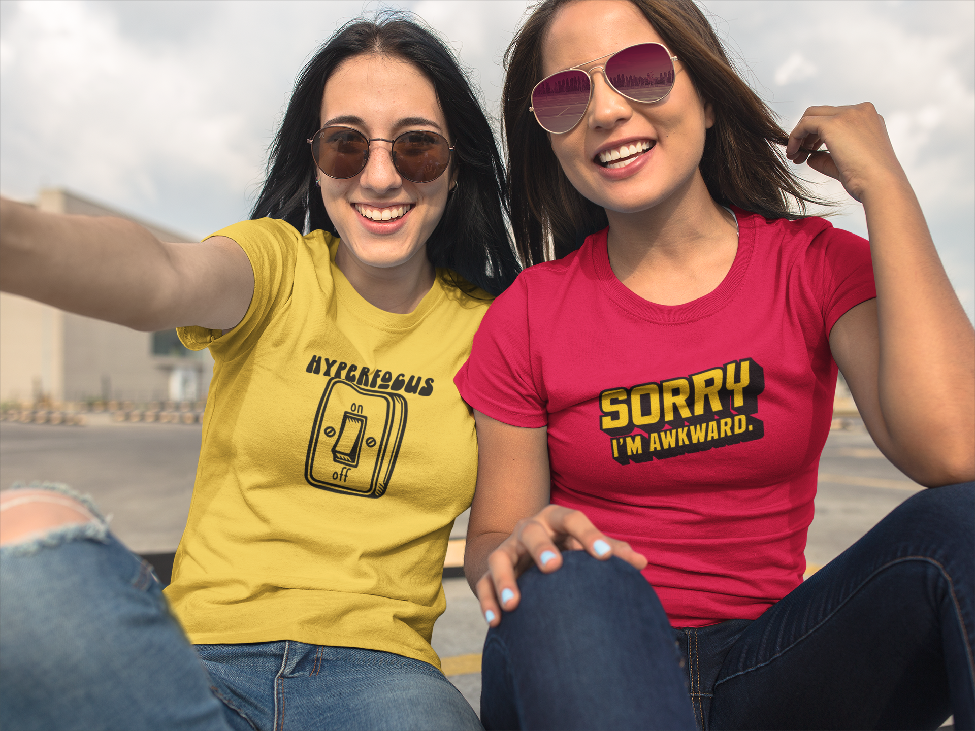 Two people sitting together, one wearing a red T-shirt and the other a yellow T-shirt, both featuring the text "Hyperfocus" with an on/off switch graphic design. Part of the Vivid Divergence Sensory Friendly Unisex Tees Range.