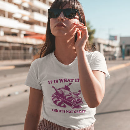 Female model wearing a white T-shirt featuring a raccoon on a skateboard with "It Is What It Is... And It's Not Great" text in pink. Part of the Vivid Divergence Sensory Friendly Unisex Tees Range.