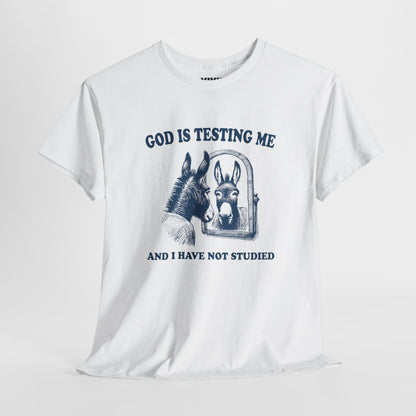 Hanging view of white T-shirt with vintage-style illustration of a distressed donkey and the phrase “God is testing me and I have not studied.” Divine pressure, zero preparation. Part of the Vivid Divergence Memewear Collection!