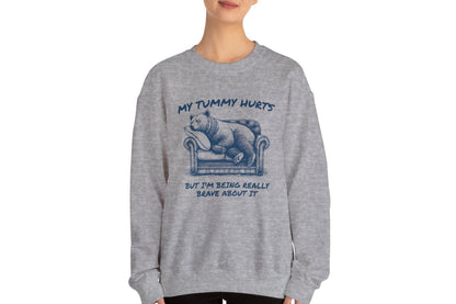 Model wearing a gray sweater jumper with the text "My Tummy Hurts But I’m Being Really Brave About It" and a cartoon illustration of a bear holding its stomach. Part of the Vivid Divergence Sensory Friendly Unisex Sweater Jumper Range.