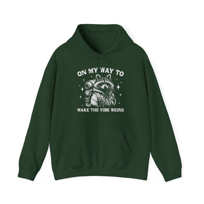 Front view of a green hoodie featuring the text "On My Way to Make the Vibe Weird" and a cartoon illustration of a raccoon holding a stick. Part of the Vivid Divergence Sensory Friendly Unisex Hoodie Range.