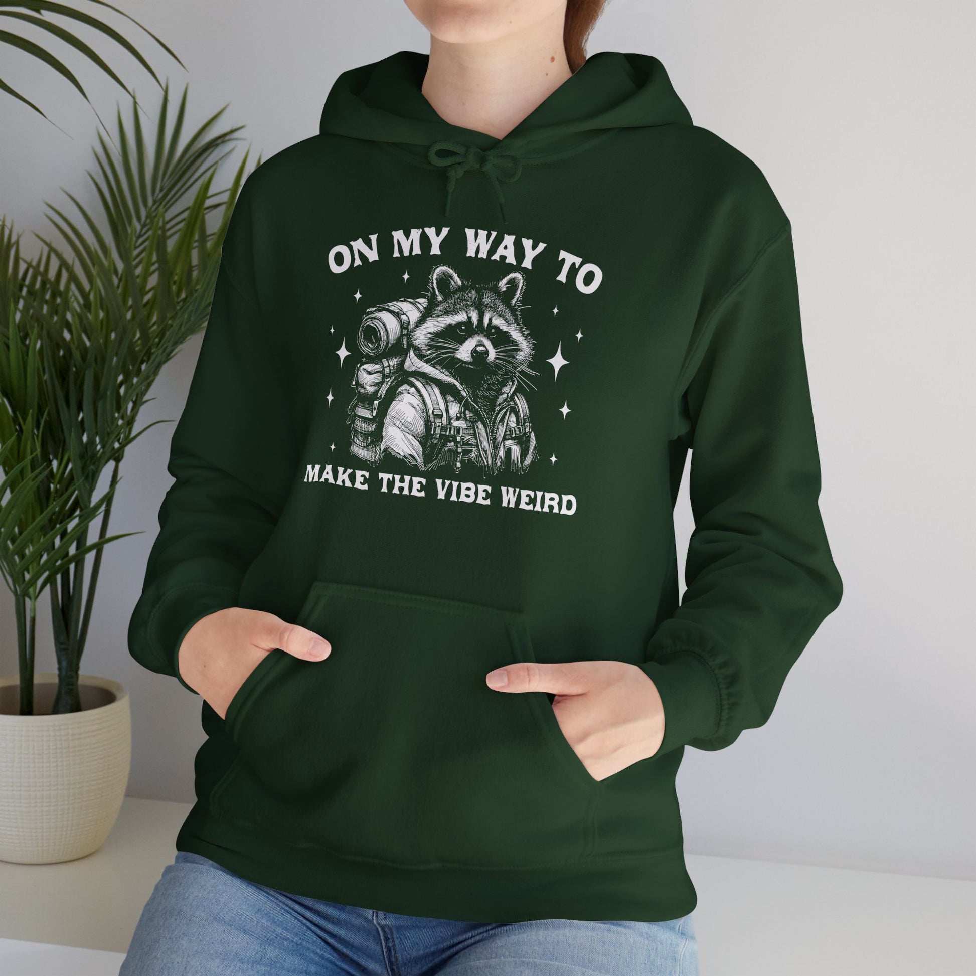 Model wearing a green hoodie featuring the text "On My Way to Make the Vibe Weird" and a cartoon illustration of a raccoon holding a stick. Part of the Vivid Divergence Sensory Friendly Unisex Hoodie Range.