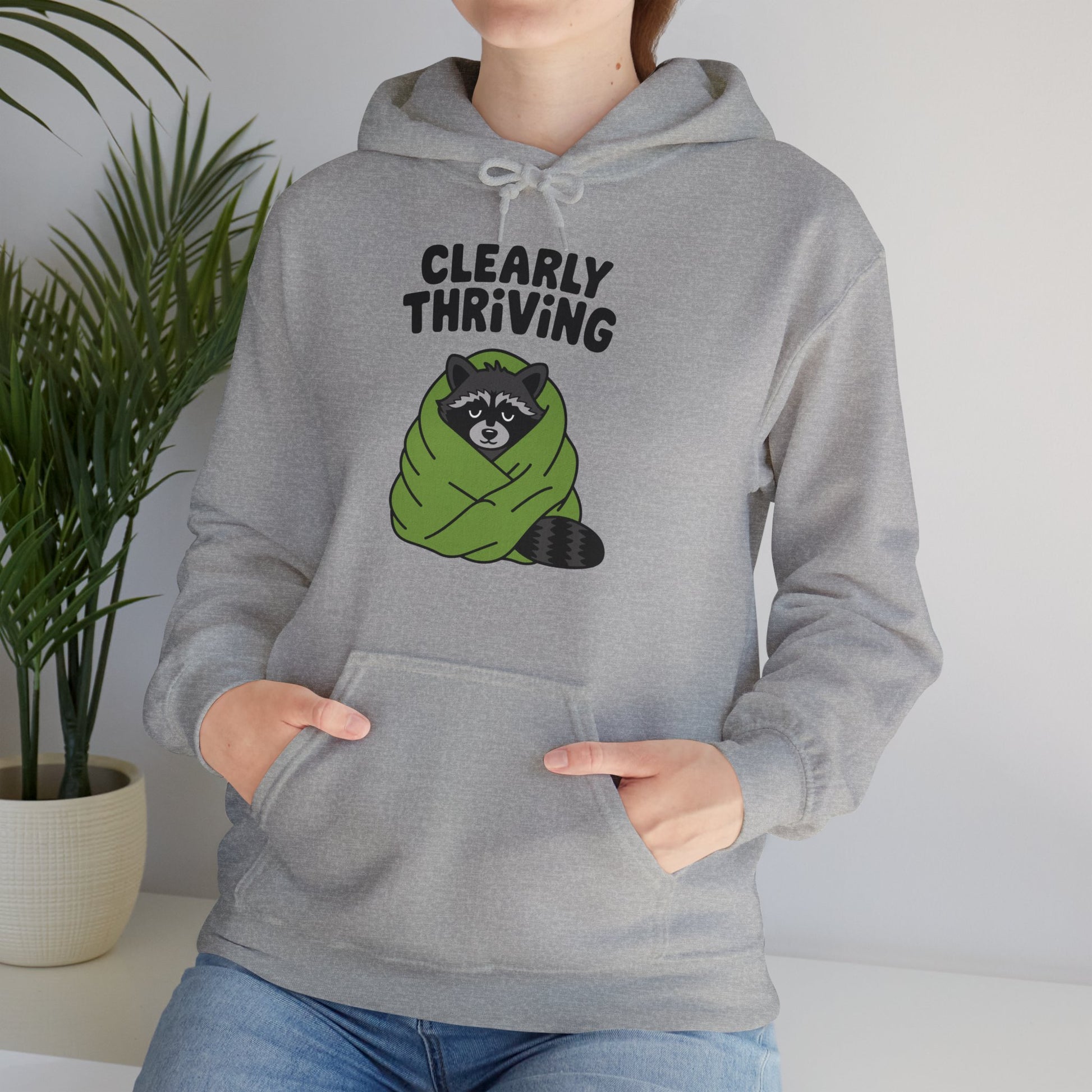 Model wearing a grey hoodie featuring the text "Clearly Thriving" and a cartoon illustration of a raccoon wrapped in a cozy blanket. Part of the Vivid Divergence Sensory Friendly Unisex Hoodie Range.