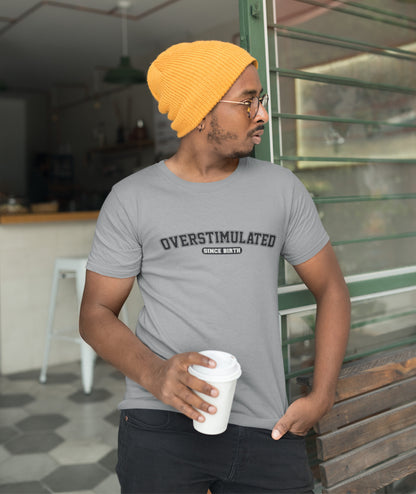 Model wearing a grey T-shirt with "Overstimulated Since Birth" in bold black text. Part of the Vivid Divergence Sensory Friendly Unisex Tees Range.