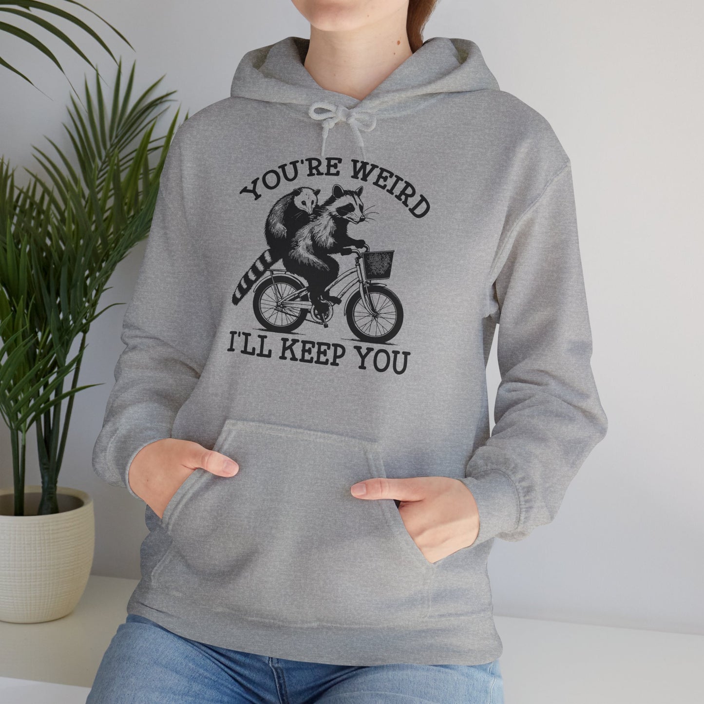Model wearing a grey hoodie featuring the text "You're Weird, I'll Keep You" with a cartoon illustration of two raccoons riding a bicycle. Part of the Vivid Divergence Sensory Friendly Unisex Hoodie Range.