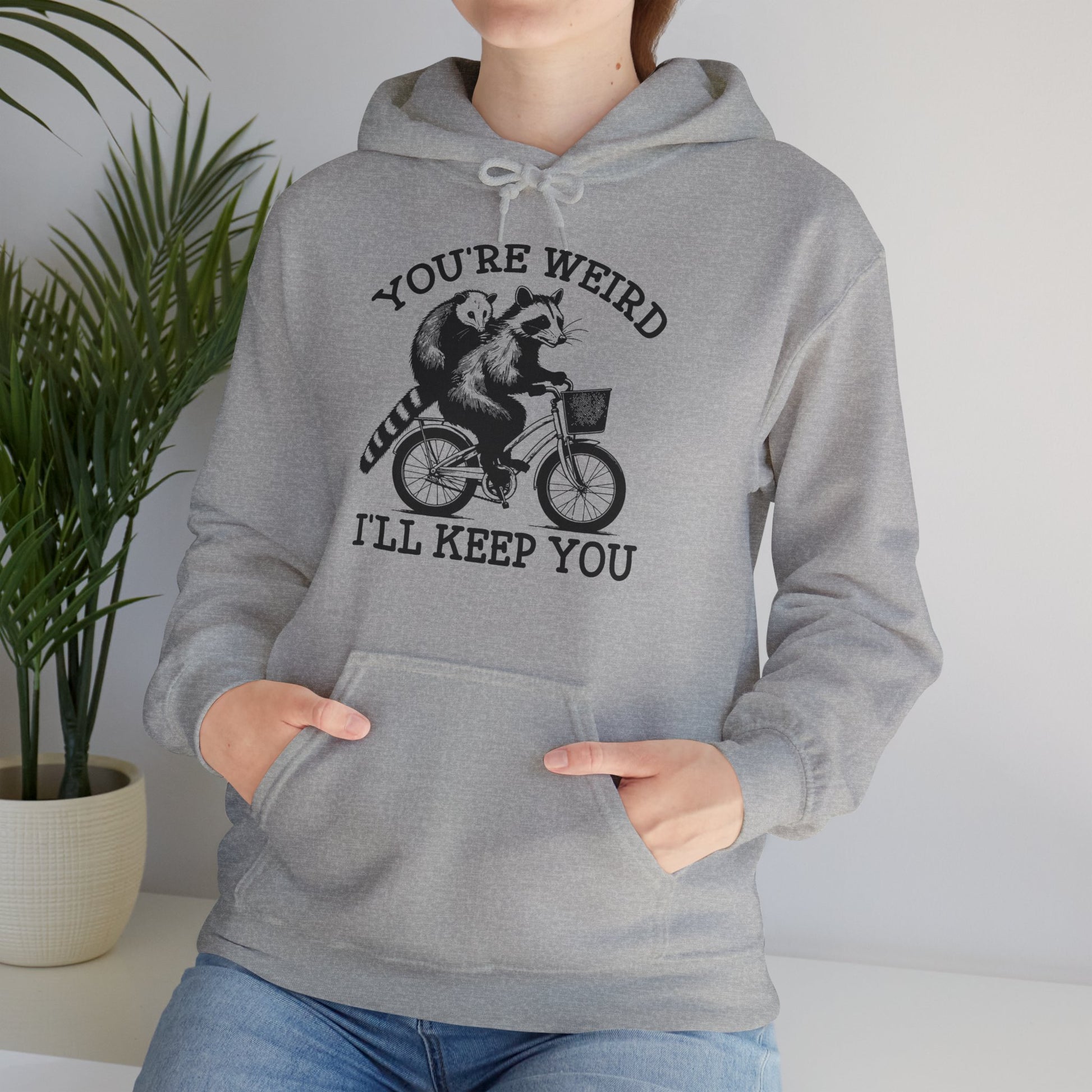 Model wearing a grey hoodie featuring the text "You're Weird, I'll Keep You" with a cartoon illustration of two raccoons riding a bicycle. Part of the Vivid Divergence Sensory Friendly Unisex Hoodie Range.