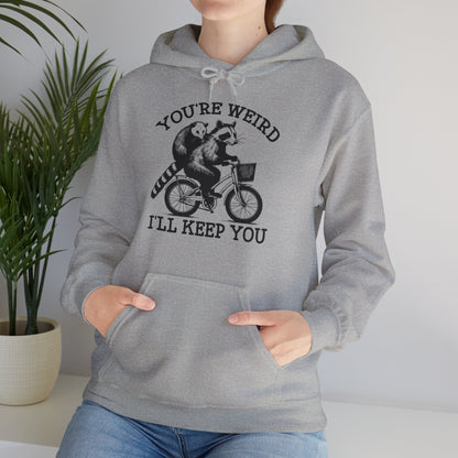 Model wearing a grey hoodie featuring the text "You're Weird, I'll Keep You" with a cartoon illustration of two raccoons riding a bicycle. Part of the Vivid Divergence Sensory Friendly Unisex Hoodie Range.