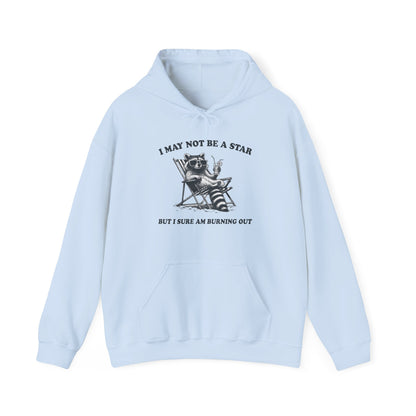 Front view of a light blue hoodie featuring a raccoon sitting in a trash can with the text "I May Not Be A Star, But I Sure Am Burning Out." Part of the Vivid Divergence Sensory Friendly Unisex Hoodie Range.