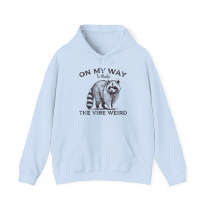 Front view of a light blue hoodie featuring the text "On My Way to Make the Vibe Weird" and a cartoon illustration of a raccoon holding a stick. Part of the Vivid Divergence Sensory Friendly Unisex Hoodie Range.