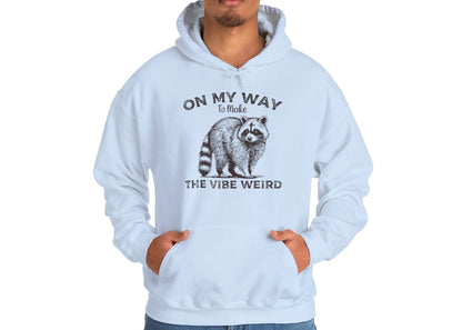 Model wearing a light blue hoodie featuring the text "On My Way to Make the Vibe Weird" and a cartoon illustration of a raccoon holding a stick. Part of the Vivid Divergence Sensory Friendly Unisex Hoodie Range.
