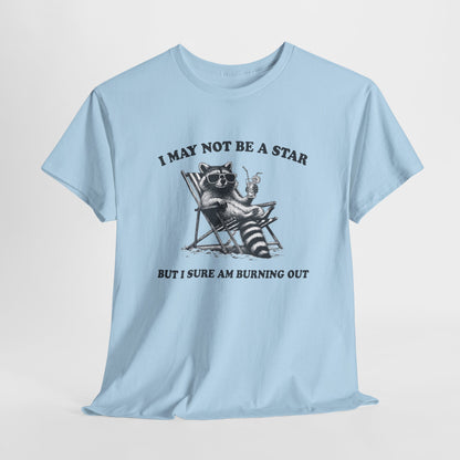 Hanging front view of a light blue T-shirt featuring a cute raccoon holding a star with text that reads "I May Not Be A Star, But I Sure Am Burning Out." Part of the Vivid Divergence Sensory Friendly Unisex Tees Range.
