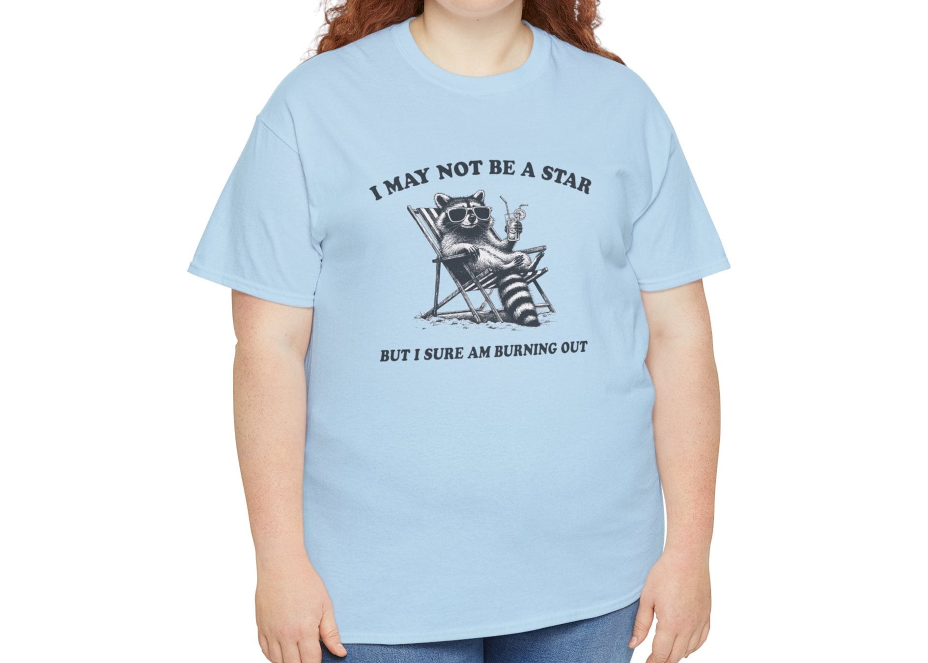 Modeled front view of a light blue T-shirt featuring a cute raccoon holding a star with text that reads "I May Not Be A Star, But I Sure Am Burning Out." Part of the Vivid Divergence Sensory Friendly Unisex Tees Range.