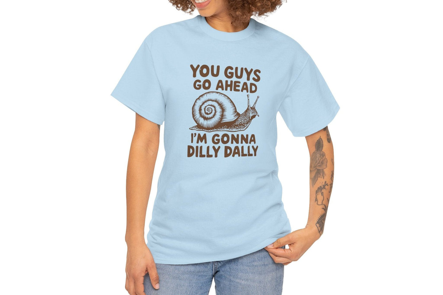 Model wearing a light blue T-shirt with the text "You Guys Go Ahead, I'm Gonna Dilly Dally" and a playful snail graphic in brown. Part of the Vivid Divergence Sensory Friendly Unisex Tees Range.