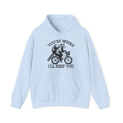 Front view of a light blue hoodie featuring the text "You're Weird, I'll Keep You" with a cartoon illustration of two raccoons riding a bicycle. Part of the Vivid Divergence Sensory Friendly Unisex Hoodie Range.