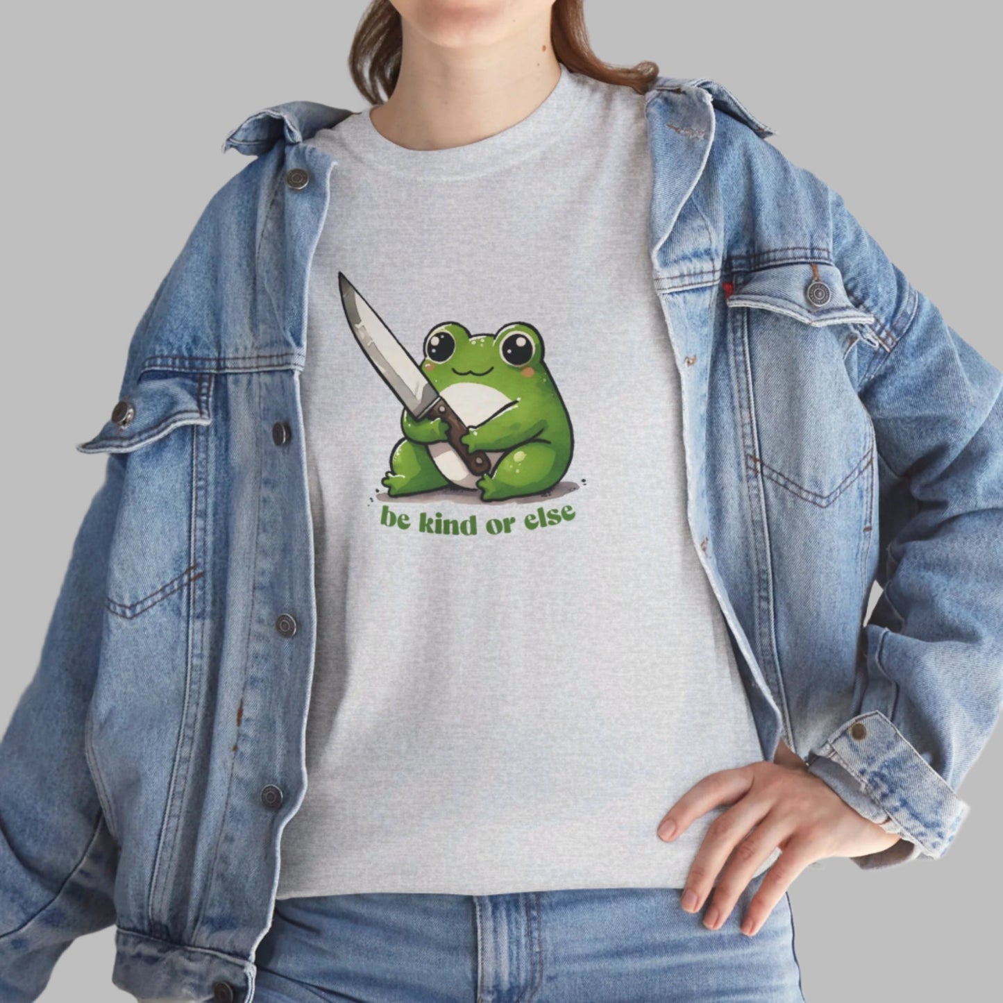 Light gray T-shirt featuring a green frog holding a knife with the text "Be Kind or Else," worn by a person styled with a denim jacket. Part of the Vivid Divergence Sensory Friendly Unisex Tees Range.