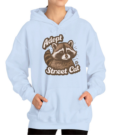Model wearing a light blue hoodie with the text "Adopt a Street Cat" and a cartoon illustration of a raccoon holding a cup. Part of the Vivid Divergence Sensory Friendly Unisex Hoodie Range.