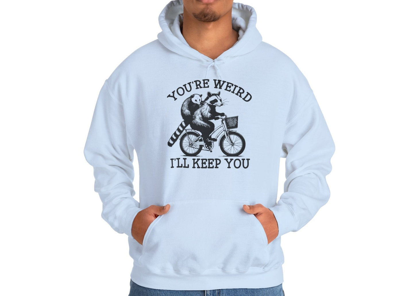 Model wearing a light blue hoodie featuring the text "You're Weird, I'll Keep You" with a cartoon illustration of two raccoons riding a bicycle. Part of the Vivid Divergence Sensory Friendly Unisex Hoodie Range.