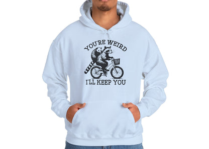 Model wearing a light blue hoodie featuring the text "You're Weird, I'll Keep You" with a cartoon illustration of two raccoons riding a bicycle. Part of the Vivid Divergence Sensory Friendly Unisex Hoodie Range.