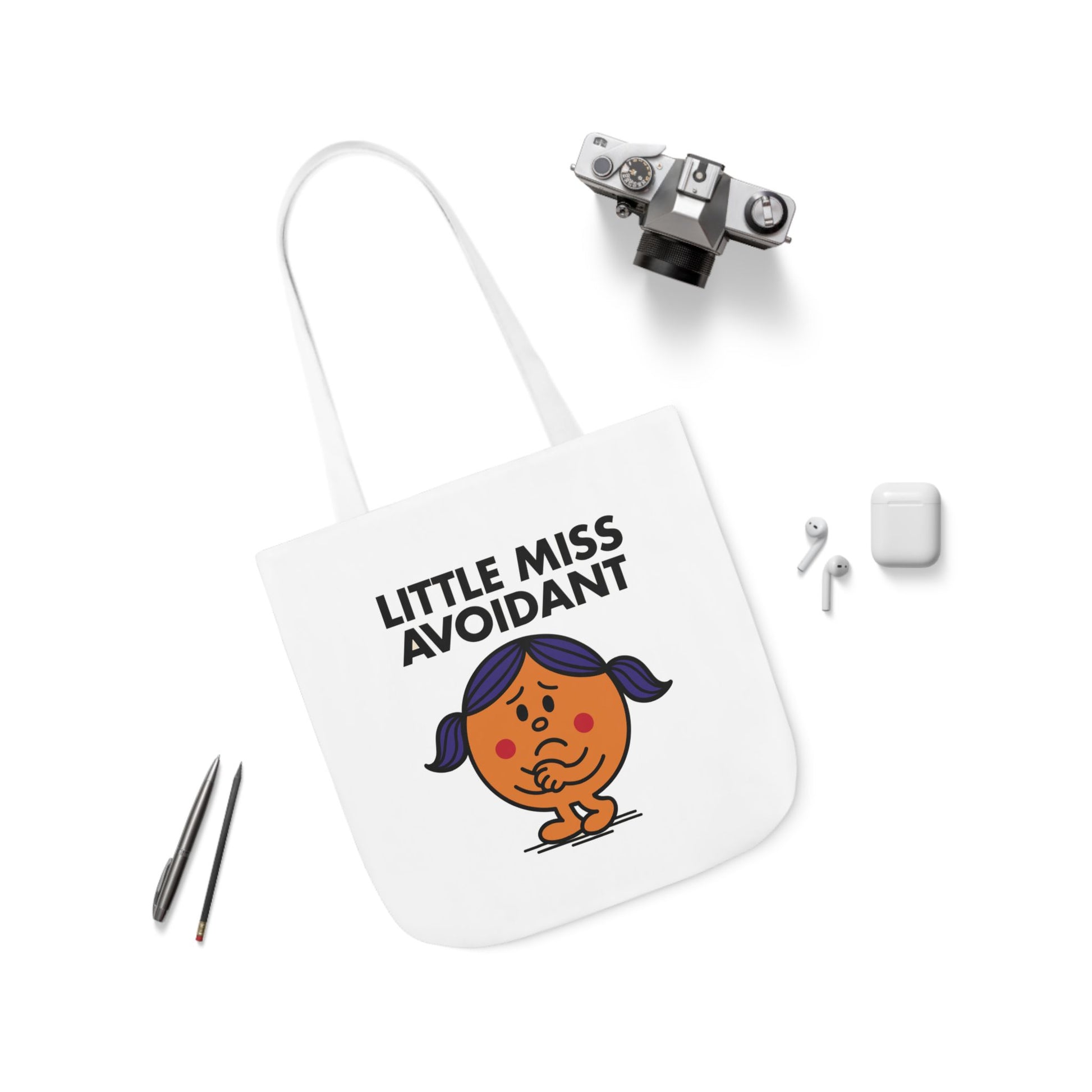 Flat lay of tote bag showing Little Miss Avoidant surrounded by pens and wireless earbuds — perfect tools for ignoring texts. Part of the Vivid Divergence Little Miss Meme Collection!
