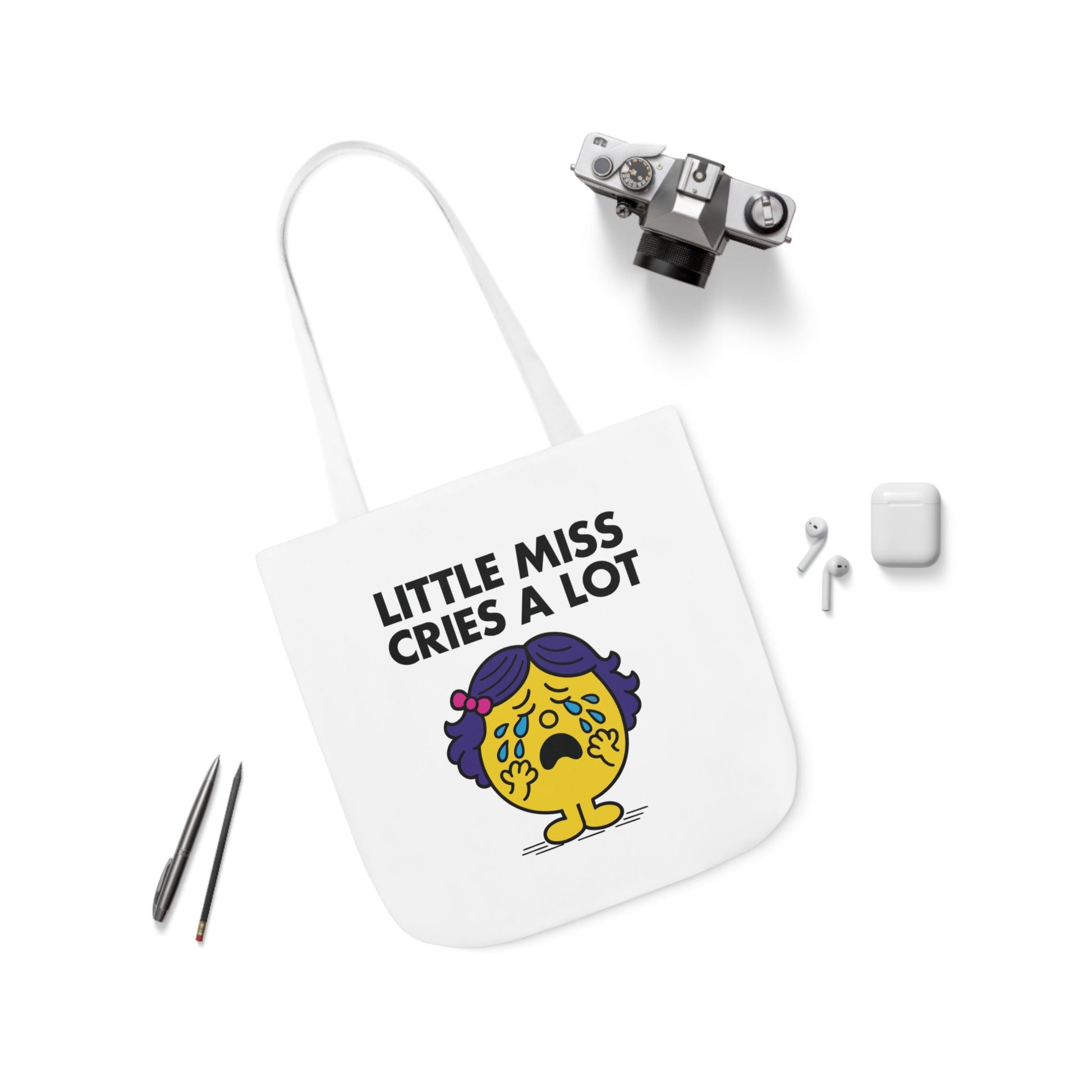 Flat lay of tote bag showing Little Miss Cries A Lot surrounded by earbuds and pens. The playlist? Sad. The vibes? Immaculate. Part of the Vivid Divergence Little Miss Meme Collection!
